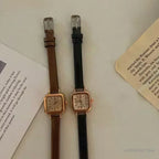 Two women's wristwatches with small square dials, one with a brown leather strap and gold case, and the other with a black leather strap and gold case.
